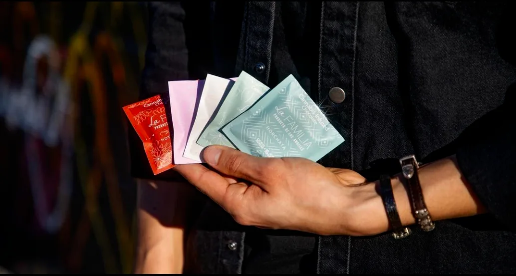 A hand holding a fanned-out set of single-serve La Familia sachets in warm Hot Chocolate Red, Fresa con Crema Pink, cream, and Horchata Blue, against a dark denim jacket in golden light.