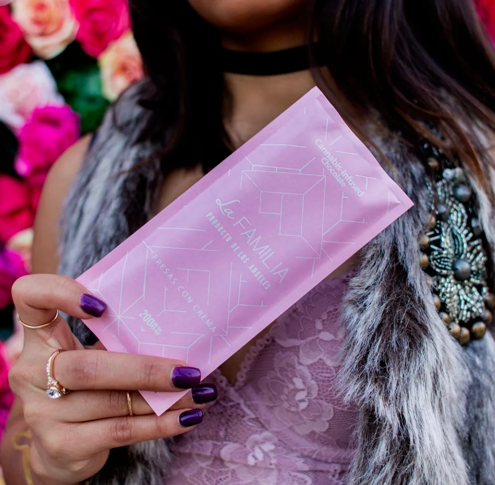 A woman with purple-painted nails holding a pink La Familia product pouch, set against a backdrop of vivid pink and magenta roses, modern, premium, and vibrant.